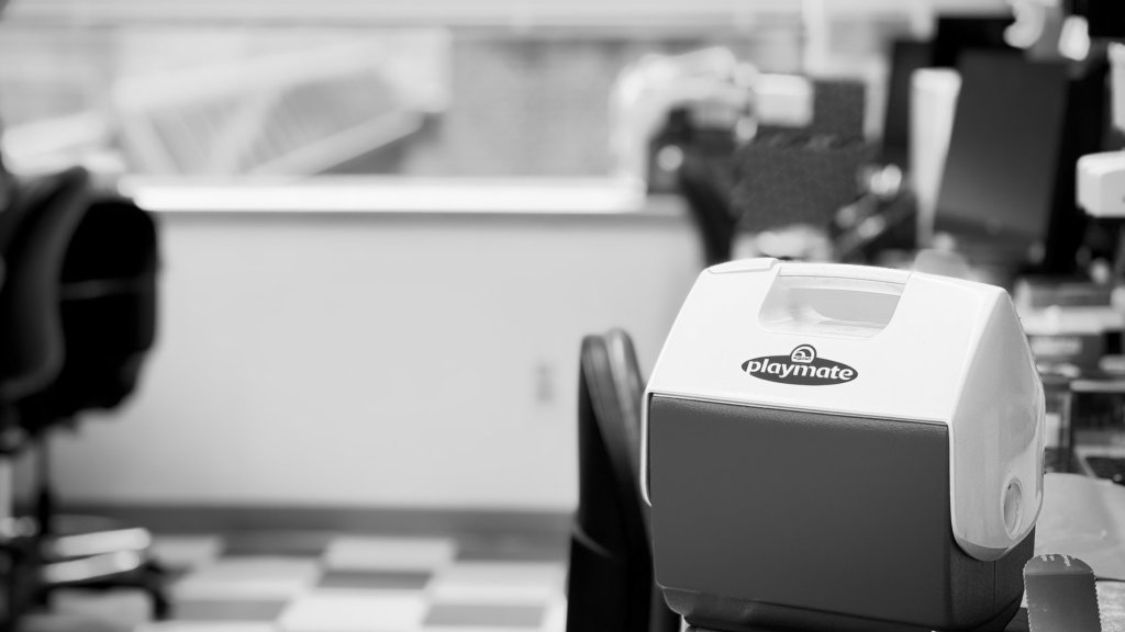 Black-and-white photograph of a small white Igloo Playmate cooler resting on a laboratory bench, with chairs, benches and equipment softly out of focus behind it.